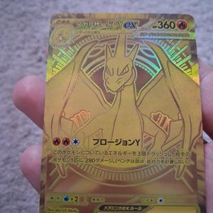 May include: A gold-colored trading card featuring a stylized illustration of a dragon-like creature. The card displays the text Mega Charizard Y ex, 360 HP, and Japanese characters throughout. It includes fire energy symbols and a 766/742 set number.