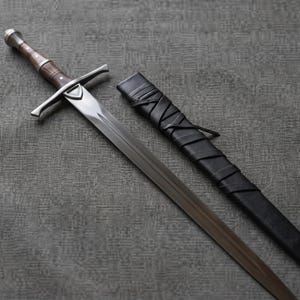 Peut inclure: Une épée en acier avec une lame à double tranchant, une garde transversale et un manche en bois brun avec des accents en métal argenté. À côté de l'épée se trouve un fourreau en cuir noir enveloppé d'un cordon en cuir, avec une pointe en métal.