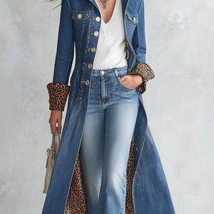 Puede incluir: Un abrigo largo de mezclilla azul con cuello y puños de estampado de leopardo, combinado con una blusa blanca de cuello en V y jeans de mezclilla azul claro. El abrigo tiene botones plateados al frente y un forro de leopardo visible por dentro.