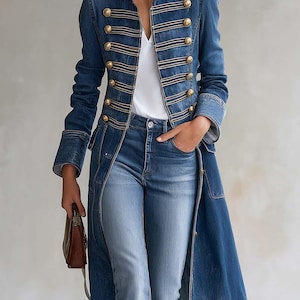 Può includere: Un lungo cappotto in denim blu con colletto alto, spalline e diverse file di passamaneria intrecciata color oro con bottoni dorati abbinati. Il cappotto è indossato sopra un top bianco e jeans blu, completato da una piccola borsa a mano marrone.
