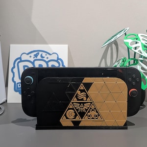May include: A black video game console dock featuring a gold-coloured triangular front plate with engraved symbols including a central crest, a crescent moon, and circular patterns, all arranged within a geometric grid of small triangles.