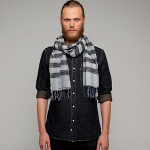 Può includere: Sciarpa a righe blu e grigie con frange. Indossata sopra una camicia di jeans scura. La camicia ha le maniche arrotolate ed è abbottonata. Lo sfondo è grigio uniforme.
