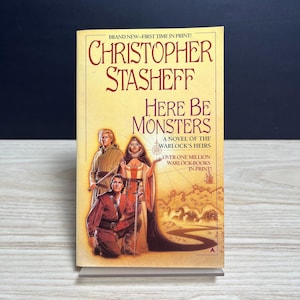 May include: A paperback book titled Here Be Monsters by Christopher Stasheff, A Novel of the Warlock's Heirs. The cover features three figures in medieval-style clothing, a sword, and a landscape illustration with a winding path and monsters.