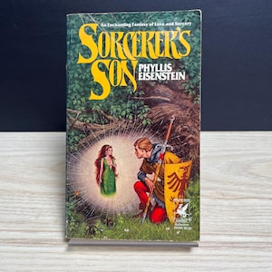 May include: A paperback book titled Sorcerer's Son by Phyllis Eisenstein. The cover features a kneeling figure in yellow and red clothing holding a sword and shield, facing a glowing female figure in a green dress within a spider web.