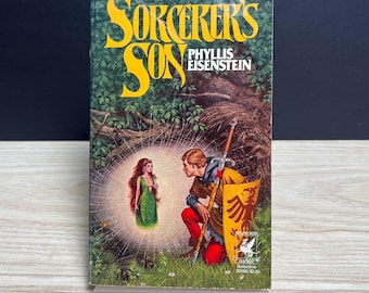 May include: A paperback book titled Sorcerer's Son by Phyllis Eisenstein. The cover features a kneeling figure in yellow and red clothing holding a sword and shield, facing a glowing female figure in a green dress within a spider web.