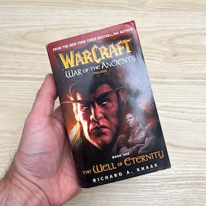 May include: A paperback book titled Warcraft War of the Ancients Trilogy Book One The Well of Eternity by Richard A. Knaak. The cover features a dark, fiery illustration of a character with glowing purple eyes and a warrior figure.