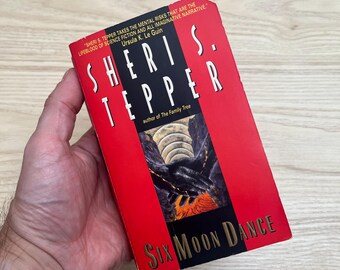 May include: A paperback book titled Six Moon Dance by Sheri S. Tepper. The cover is primarily red with a black central panel featuring an illustration of dark, textured hands reaching toward glowing orange light and circular moon-like shapes.