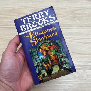 May include: A mass market paperback book titled The Elfstones of Shannara by Terry Brooks. The dark blue cover features an illustration of three fantasy characters standing before a stone archway. The Del Rey logo is visible at the bottom.