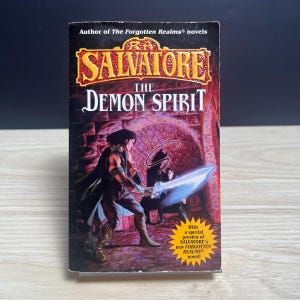 May include: A paperback book titled The Demon Spirit by R.A. Salvatore. The cover features a dark fantasy illustration of a figure holding a glowing blue sword in a stone chamber. A yellow starburst graphic on the cover mentions a special preview.