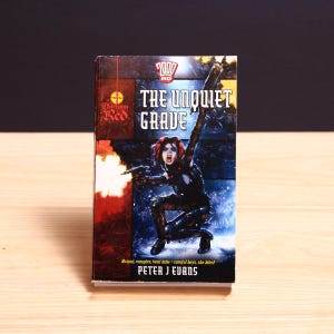 May include: A paperback book titled The Unquiet Grave by Peter J Evans, featuring a 2000 AD logo and Durham Red branding. The cover art depicts a character with red hair and fangs, crouching while firing a weapon, with the text Mutant, vampire, total babe - careful boys, she bites! printed at the bottom.