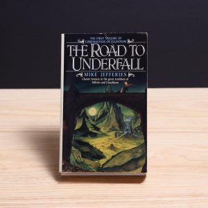 May include: A paperback book titled The Road to Underfall by Mike Jefferies, the first volume of Loremasters of Elundium. The cover features a dark archway framing a landscape with mountains, a full moon, and a structure under a night sky.