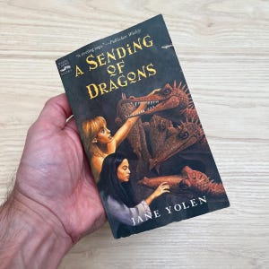 May include: A paperback book titled A Sending of Dragons by Jane Yolen. The black cover features an illustration of two people interacting with three brown, textured dragons. The title is printed in large, gold, serif lettering.