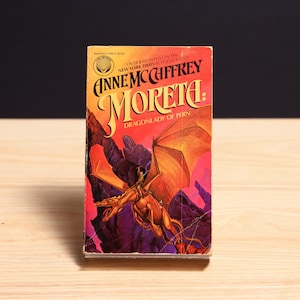 May include: A mass market paperback book titled Moreta: Dragonlady of Pern by Anne McCaffrey. The cover features a golden dragon with a rider against a backdrop of dark purple mountains and a vibrant orange sky, with text at the top.
