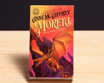 May include: A mass market paperback book titled Moreta: Dragonlady of Pern by Anne McCaffrey. The cover features a golden dragon with a rider against a backdrop of dark purple mountains and a vibrant orange sky, with text at the top.