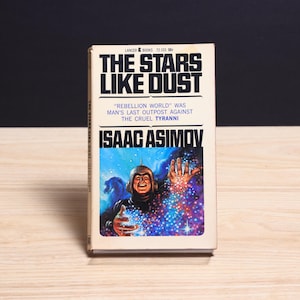 Könnte beinhalten: Ein Vintage-Taschenbuch mit dem Titel The Stars Like Dust von Isaac Asimov, herausgegeben von Lancer Books. Das Cover zeigt eine farbenfrohe Illustration einer Figur mit metallischem Helm, umgeben von leuchtendem Sternenstaub und Licht.
