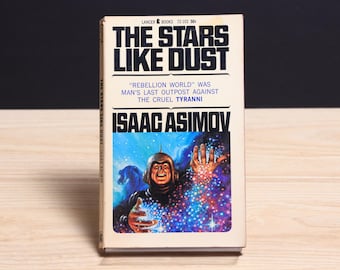May include: A vintage paperback book titled The Stars Like Dust by Isaac Asimov, published by Lancer Books. The cover features a colorful illustration of a figure in a metallic helmet surrounded by glowing, multi-colored stardust and cosmic light.