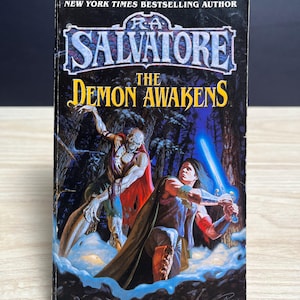 May include: A mass market paperback book titled The Demon Awakens by R.A. Salvatore. The cover features a dark forest scene with a pale, undead creature in a red cloak and a warrior wielding a glowing blue sword, set against a black background.