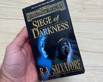 Puede incluir: Un libro de bolsillo titulado Siege of Darkness de R.A. Salvatore, parte de la serie Forgotten Realms. La portada negra presenta letras doradas y una ilustración de dos figuras con efectos de iluminación en tonos azules y verdes.