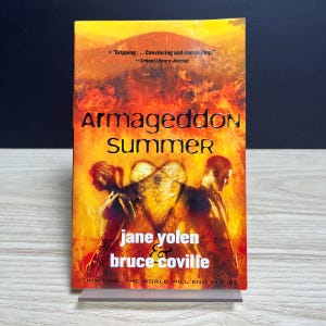 May include: A paperback book titled Armageddon Summer by Jane Yolen and Bruce Coville. The cover features a fiery orange and yellow background with silhouettes of two people standing back-to-back, separated by a glowing heart-shaped light.