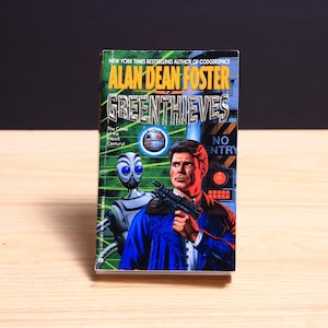 May include: A paperback book titled Greenthieves by Alan Dean Foster. The cover features a man in a blue jacket holding a black firearm, a silver robot with large blue eyes, a floating drone, and a sign that reads NO ENTRY in yellow and black.