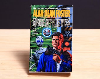 May include: A paperback book titled Greenthieves by Alan Dean Foster. The cover features a man in a blue jacket holding a black firearm, a silver robot with large blue eyes, a floating drone, and a sign that reads NO ENTRY in yellow and black.