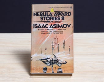 Może przedstawiać: Książka w miękkiej oprawie pod tytułem Nebula Award Stories 8, pod redakcją Isaaca Asimova. Okładka przedstawia surrealistyczny, beżowy krajobraz z abstrakcyjnymi, metalicznymi i smukłymi postaciami. Tekst zawiera tytuł oraz nazwiska autorów.