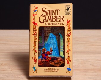 May include: A paperback book titled Saint Camber by Katherine Kurtz, Volume II in the Legends of Camber of Culdi. The cover features a tan background with red and gold floral borders and an illustration of a person in red and blue clothing.