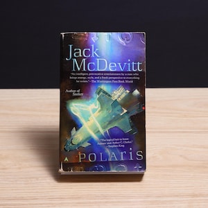 May include: A paperback book titled Polaris by Jack McDevitt. The cover features a blue and green spaceship illustration against a dark, cosmic background with glowing light effects. Text includes author name, title, and a quote by Stephen King.
