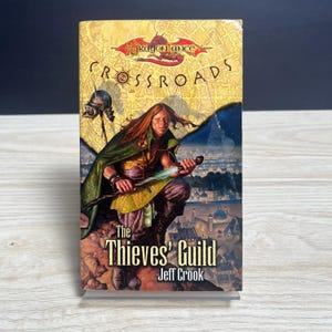 May include: A paperback book titled Dragonlance Crossroads: The Thieves' Guild by Jeff Crook. The cover features a fantasy illustration of a long-haired figure in a green cloak holding a glowing sword, with a city skyline and a dragon emblem above.