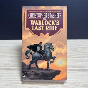 May include: A paperback book titled The Warlock's Last Ride by Christopher Stasheff. The cover features an illustration of a person in a dark cape riding a metallic, mechanical horse on a cliffside against a sunset sky with a large planet.