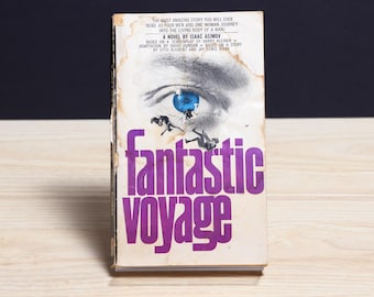 May include: A paperback book titled Fantastic Voyage by Isaac Asimov. The cover features a large blue eye, silhouettes of people, and text detailing the story of four men and one woman journeying into a human body. The edges show wear and aging.