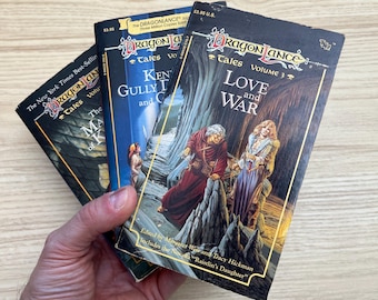 May include: Three Dragonlance Tales paperback books featuring fantasy cover art. The top book is Volume 3, Love and War, edited by Margaret Weis and Tracy Hickman. The covers display illustrations of characters in robes and armor with gold text.