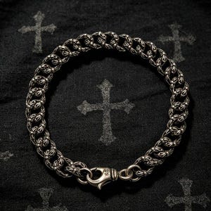Könnte beinhalten: Ein silbernes Kettenarmband mit ineinandergreifenden Gliedern, die mit kreuzförmigen Gravuren versehen sind. Das Armband wird mit einem Karabinerverschluss geschlossen, der kleine Gravuren auf der Oberfläche aufweist.