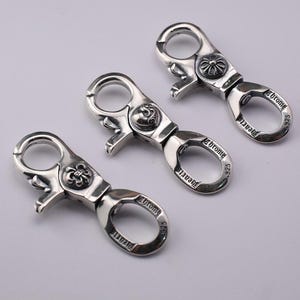 Könnte beinhalten: Drei silberfarbene Karabinerverschlüsse aus Metall mit Drehmechanismus. Jeder Verschluss zeigt ein kreisförmiges Emblem und die eingravierte Aufschrift Chrome Hearts 925. Die Beschläge haben eine polierte Oberfläche und einen Federverschluss.