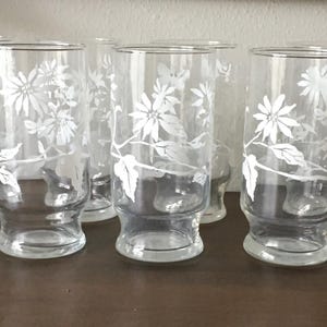 May include: Set of six clear glass drinking glasses with a white floral design. The glasses have a wide base and a tall, cylindrical shape.