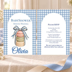 Puede incluir: Una tarjeta de invitación para baby shower con un borde de cuadros vichy azul y blanco y un arco central. El diseño incluye una pila de tres macarons atados con una cinta a rayas azules y blancas, el nombre Olivia y los detalles.