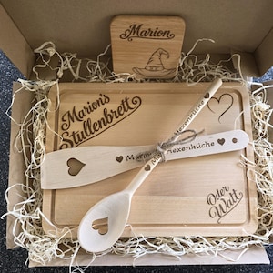 Könnte beinhalten: Ein Küchenset aus Holz in einem Karton mit einem rechteckigen Schneidebrett mit der Gravur "Marion's Stullenbrett" und "Oder Watt", einem Holzlöffel und Pfannenwender mit Herz-Ausschnitten sowie einem kleinen quadratischen Schild mit Hexenhut.