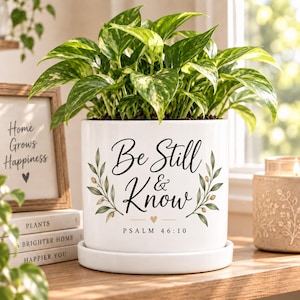 May include: A white ceramic cylindrical plant pot with a matching saucer, featuring the black printed text Be Still & Know Psalm 46:10, flanked by green olive branch illustrations and a small gold heart, containing a variegated green pothos plant.