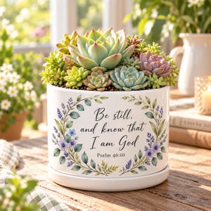 May include: A white ceramic planter with a matching saucer, filled with a variety of green and pink succulents. The front of the planter features a floral wreath design surrounding the black text: Be still, and know that I am God, Psalm 46:10.