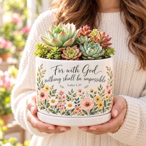 May include: A white ceramic planter featuring a floral design with pink, yellow, and white flowers. The text reads, "For with God nothing shall be impossible Luke 1:37." The planter is filled with a variety of green and pink-tipped succulent plants.