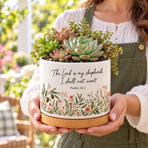 May include: A white ceramic planter with a wooden base containing various green and pink succulents. The planter features the black text "The Lord is my shepherd; I shall not want Psalm 23:1" above a colorful botanical illustration of wildflowers.