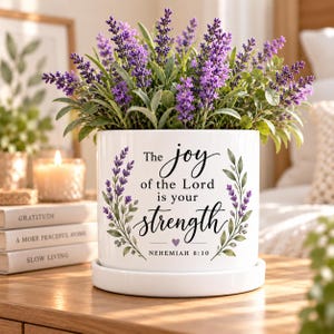 May include: A white ceramic cylindrical planter with a matching saucer, featuring a purple lavender sprig design on both sides and the black printed text: The joy of the Lord is your strength, NEHEMIAH 8:10, with a small purple heart icon.