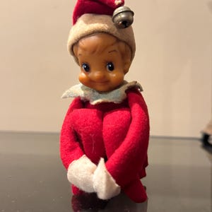 May include: A small elf figurine with a tan face, rosy cheeks, and a smiling expression. The elf wears a red and beige felt hat topped with a silver jingle bell, a red felt suit with white cuffs, and a white collar around the neck.