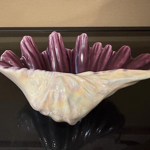 May include: A ceramic bowl shaped like a large open clam shell. The exterior features a glossy, iridescent cream and pale yellow finish with a textured, rippled surface. The interior is a deep, solid plum purple with prominent vertical ridges.