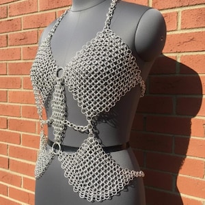 Puede incluir: Un top halter de cota de malla plateada construido con anillos de metal entrelazados. La prenda presenta un corpiño triangular conectado por una tira de cadena vertical central a una sección inferior en la cintura, con tirantes finos.