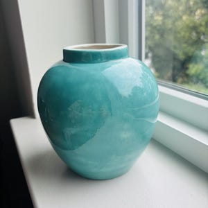 May include: A small, round ceramic vase with a glossy, vibrant turquoise glaze. The vase features a short, narrow neck and a wide, bulbous body with subtle variations in the blue-green color, creating a smooth, reflective surface finish.