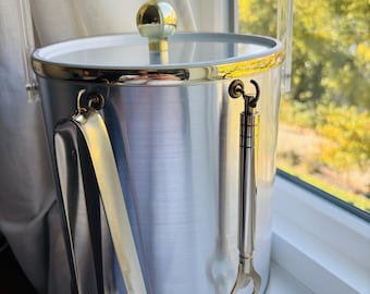 Könnte beinhalten: Ein zylindrischer Eiswürfelbehälter aus gebürstetem Silber mit goldfarbenem Rand und Deckelknopf. Er verfügt über einen schwenkbaren Griff aus klarem Acryl sowie eine silberfarbene Metall-Eiszange und einen passenden Flaschenöffner an der Seite.