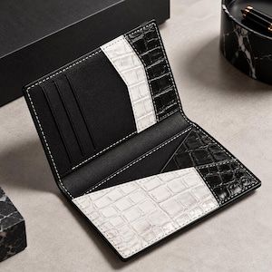Könnte beinhalten: Ein aufklappbares Portemonnaie mit einem kontrastierenden Design aus schwarz-weiß geprägten Krokodilleder-Einsätzen. Das Innere verfügt über mehrere Kartenfächer und ein Mittelfach, alles mit weißen Nähten an den Kanten versehen.