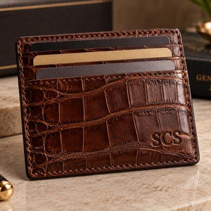Peut inclure: Un porte-cartes en cuir marron avec un motif texturé en peau de crocodile. L'accessoire rectangulaire comporte trois fentes horizontales contenant des cartes noires et beiges. Le coin inférieur droit est gaufré avec les lettres SCS.