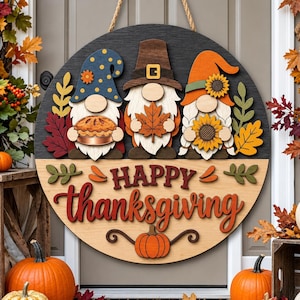 Può includere: Una targa circolare in legno per porta con tre gnomi con cappelli autunnali, che tengono in mano una torta, una foglia d'acero e un girasole. La targa ha una parte superiore scura e una chiara con la scritta HAPPY thanksgiving.